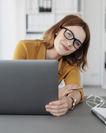 Amused young businesswoman peering around her laptop at the camera with a mischievous smile as she works at her desk in the office